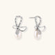 Silver bow-shaped stud earrings with baguette crystals and dangling white freshwater pearls on cream background